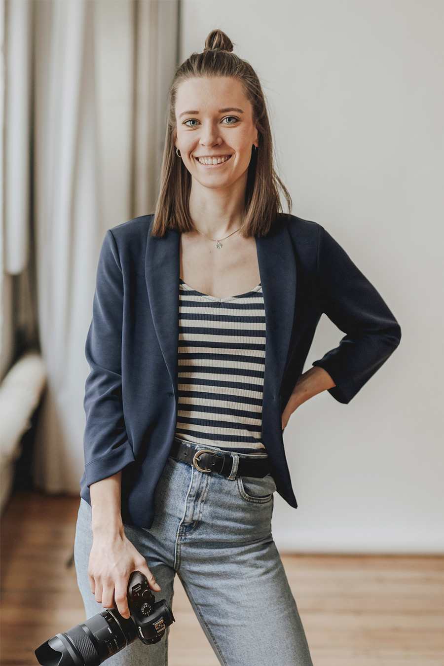 Portrait von Sarah Sembritzki. Sarah steht mit einer Kamera in der rechten Hand und lächelt in die Kamera. Der Hintergrund ist weiß. Man erkennt ein Fenster. Sarah trägt einen blauen Blazer und ein gestreiftes Top, dazu eine hellblaue Jeans.